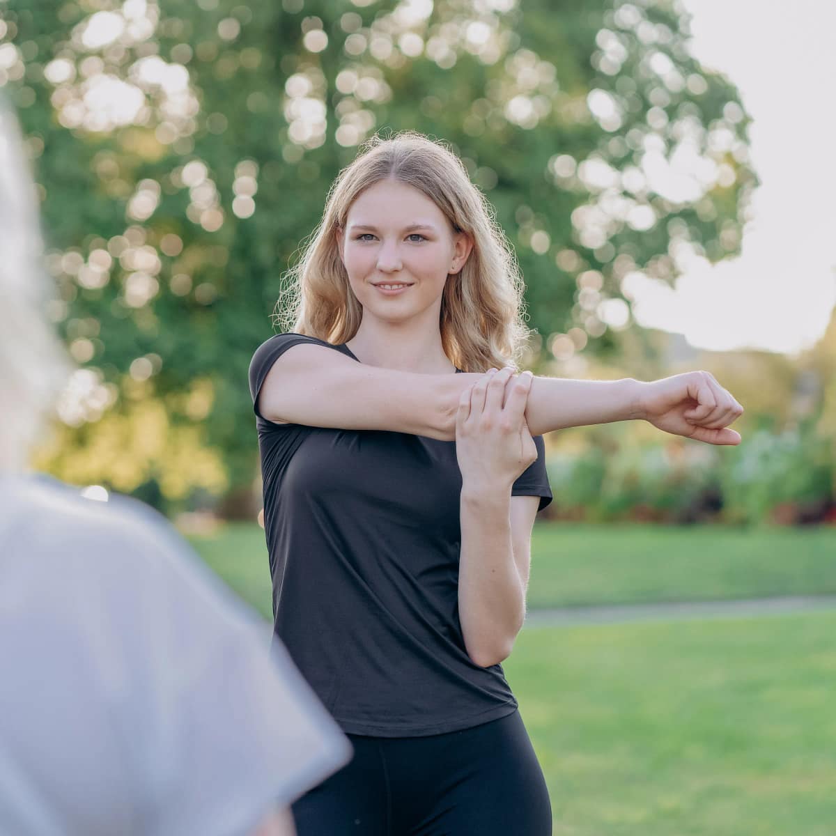 Personal Trainerin Lara Kölzow dehnt im Park ihre Schulter und lächelt in die Kamera