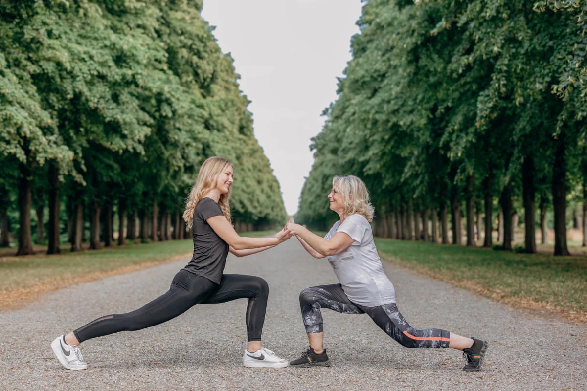 Personal Trainerin Lara Kölzow und eine Kundin im Ausfallschritt, die sich auf einem Weg in einer Baumallee an den Händen halten