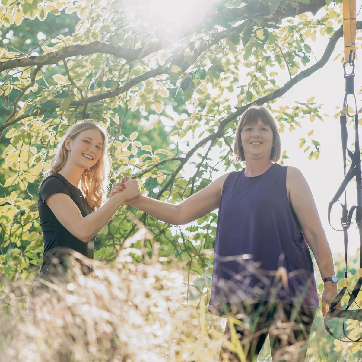 Personal Trainerin Lara Kölzow und eine Kundin geben sich lachend die Hand unter einem Baum im Gegenlicht, daneben hängt ein Schlingentrainer